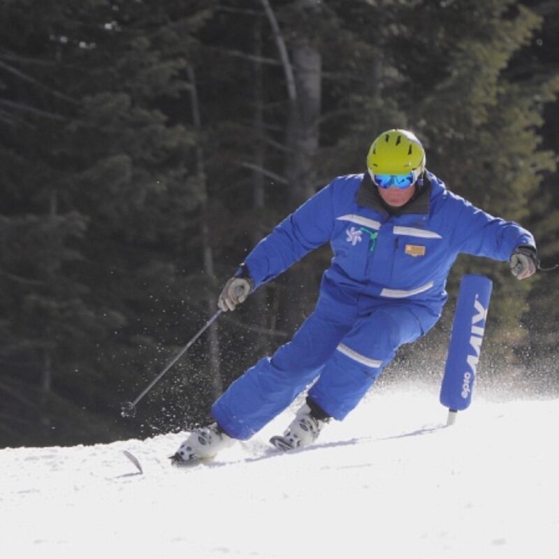 Coach Jon carving past a race gate in ski school uniform and helmet