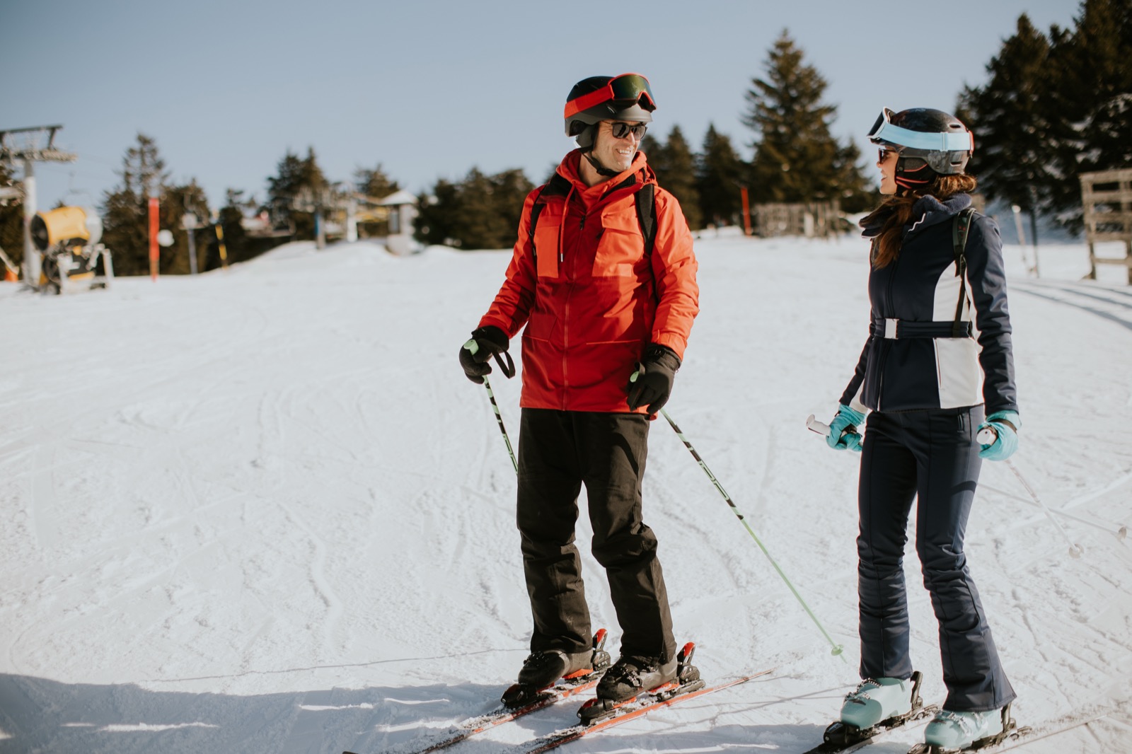 Two skiers wearing helmets standing on a groomed run with a chairlift behind them