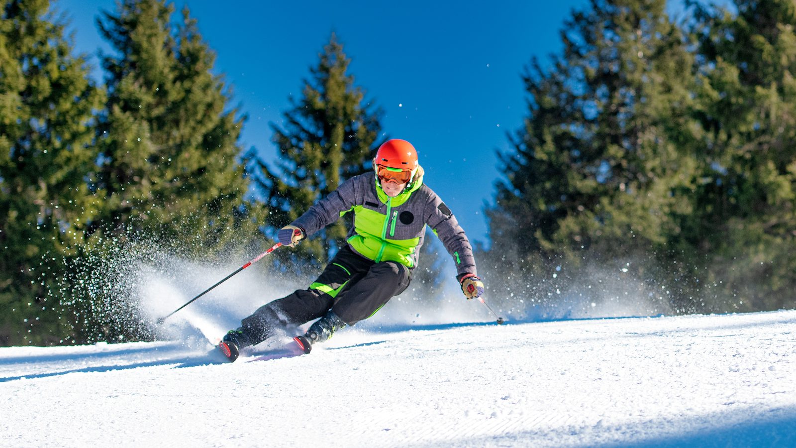 A skier in an orange helmet carving a turn on a groomed run, edge angle and snow spray visible
